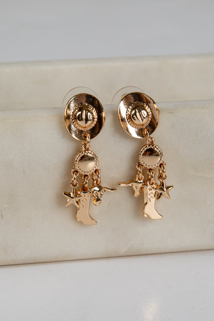 Gold earrings with cowboy boot and star charms on a beige surface.