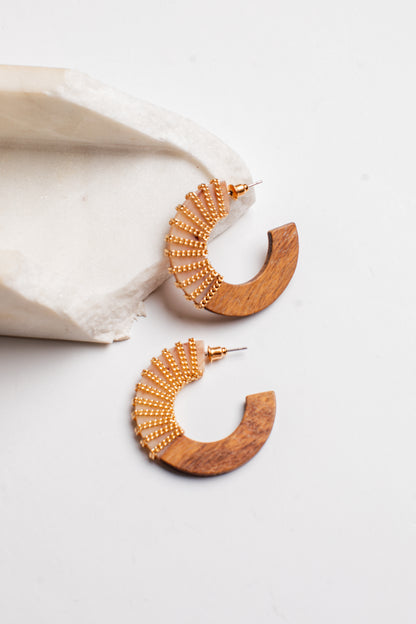 A pair of gold and wood crescent-shaped hoop earrings displayed on a clean white surface next to a textured white stone prop.