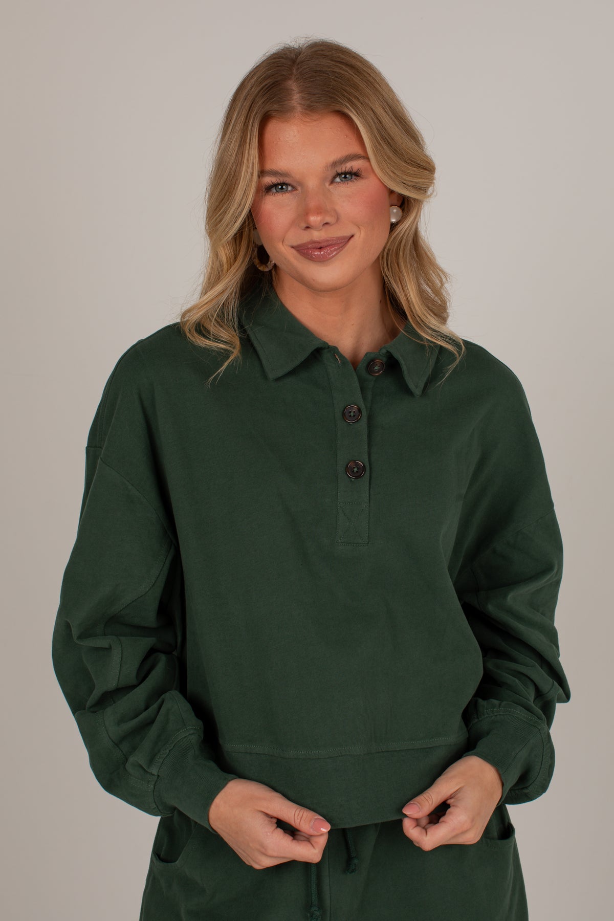 A woman modeling a dark forest green long-sleeve polo shirt with a buttoned collar, set against a plain light gray studio background.