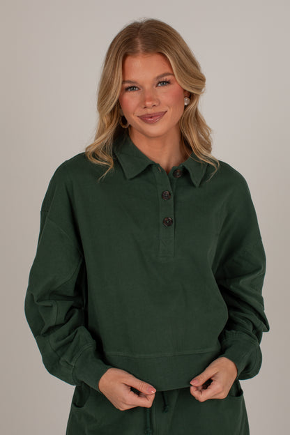 A woman modeling a dark forest green long-sleeve polo shirt with a buttoned collar, set against a plain light gray studio background.