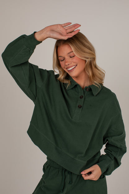 A woman wearing a dark forest green long-sleeve collared pullover top with a button placket, posing with one hand on her head against a solid light gray background.
