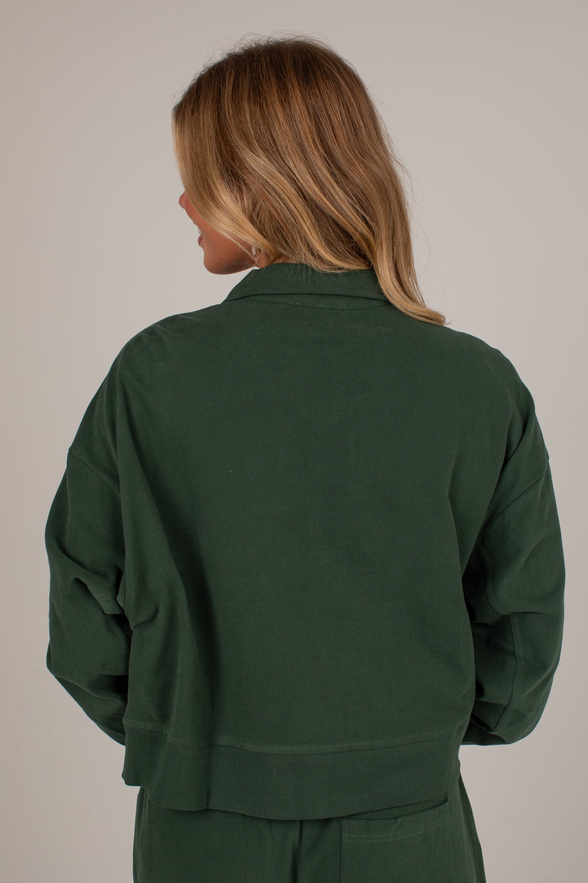 A woman shown from the back wearing a dark forest green cropped jacket and matching green pants against a plain light gray studio background.