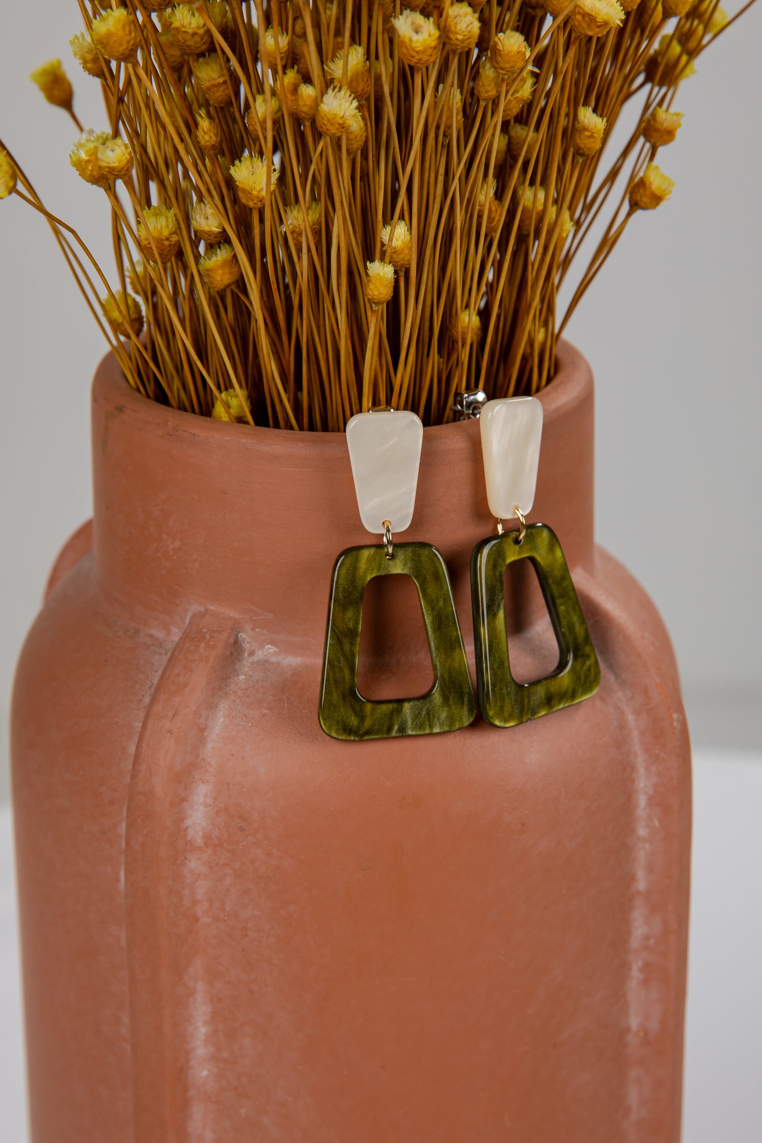 Green earrings displayed in a terracotta vase with dried flowers.