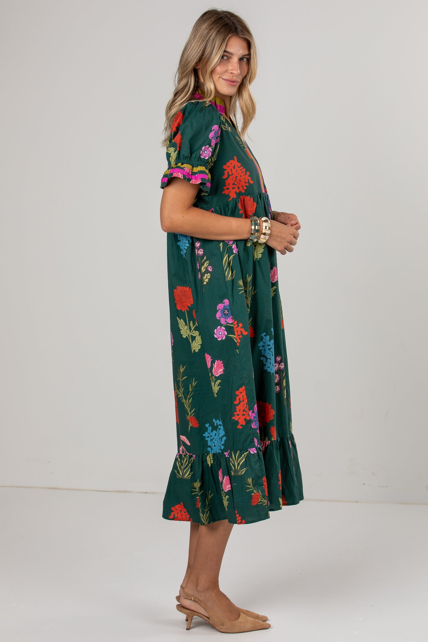A woman modeling a dark green midi dress with a colorful floral print, featuring short puff sleeves with ruffled trim and a tiered ruffle hem, paired with tan heels against a plain light gray studio background.