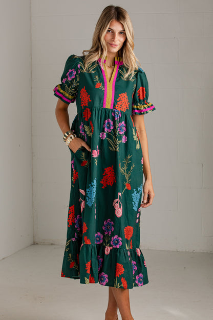 A woman modeling a dark green midi dress with a vibrant multicolor floral print, short puff sleeves, and a pink trim along the split neckline, standing in a white studio corner.