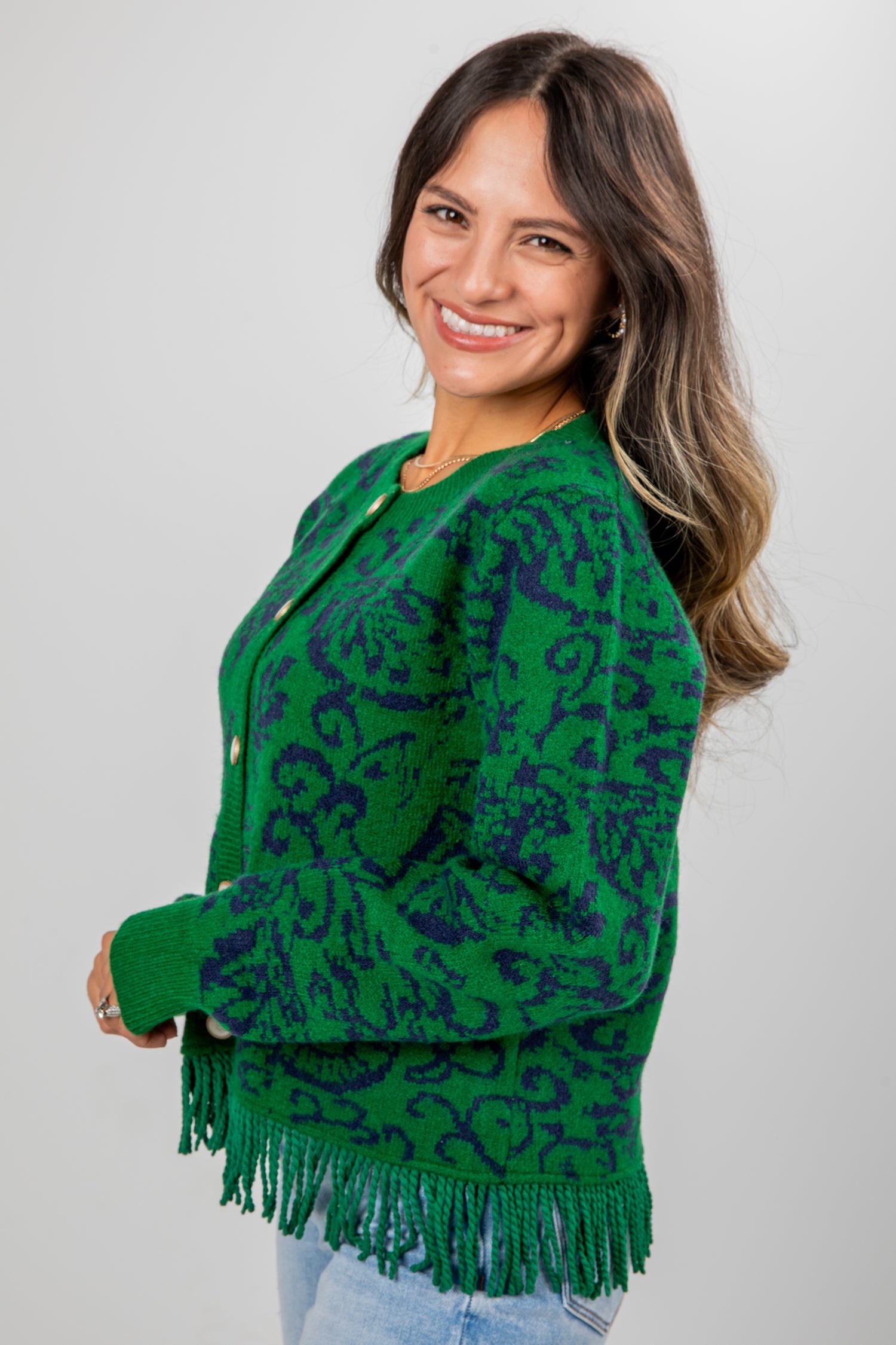 Woman wearing a green patterned cardigan with fringe on a plain background