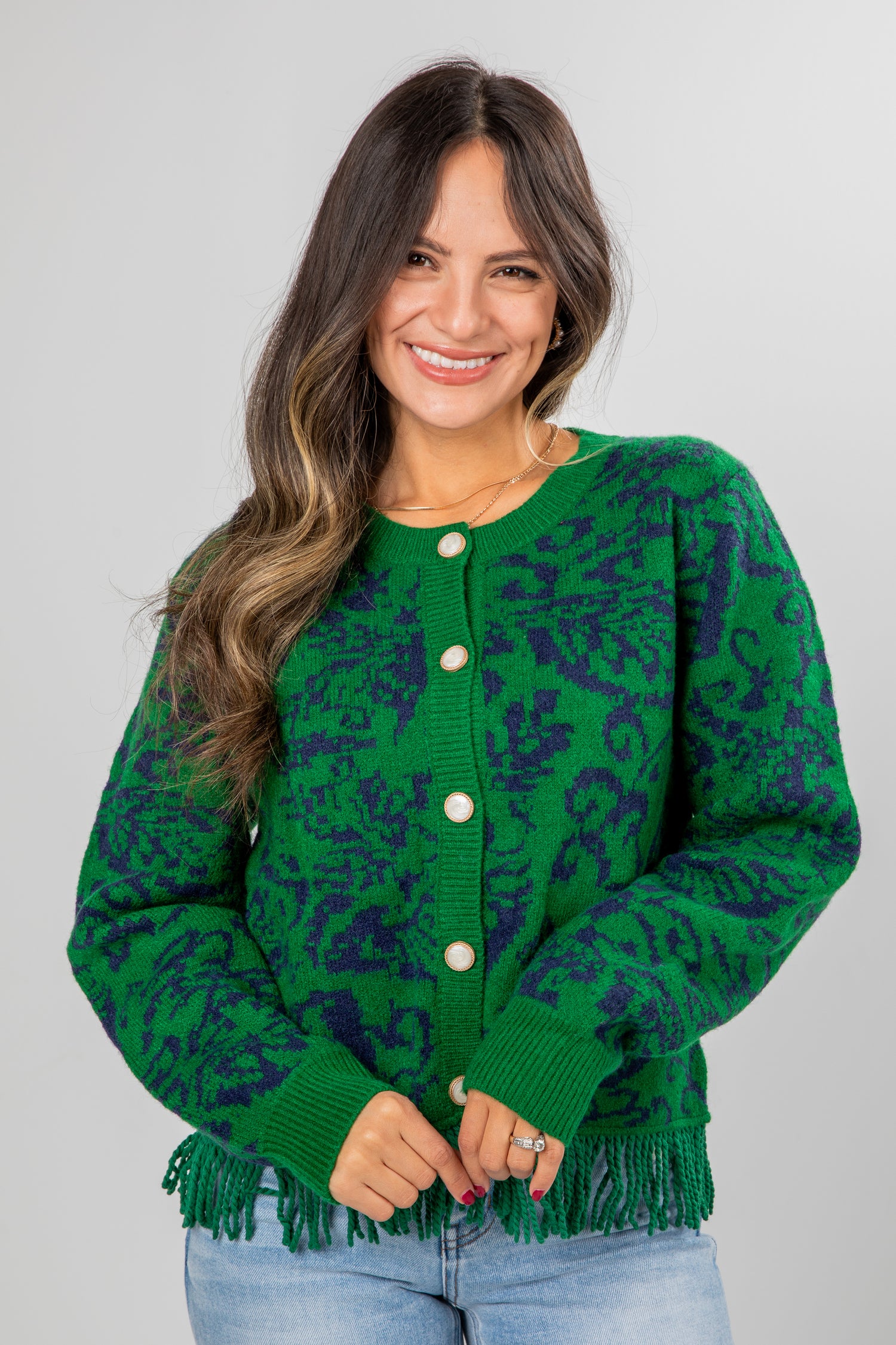 Woman wearing a green patterned cardigan with fringe on a plain background
