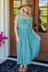 A woman wearing a green and white gingham midi dress, a straw hat, and sandals stands on a black doormat in front of a brown wooden door with glass panes.