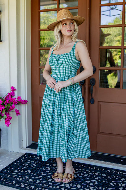 A woman wearing a green and white gingham midi dress, a straw hat, and tan sandals stands on a black patterned doormat in front of a brown front door with pink flowers on the left.