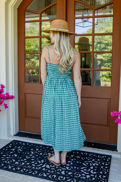 A woman stands with her back to the camera in front of a brown front door, wearing a green and white gingham midi dress, a straw hat, and sandals on a black doormat.