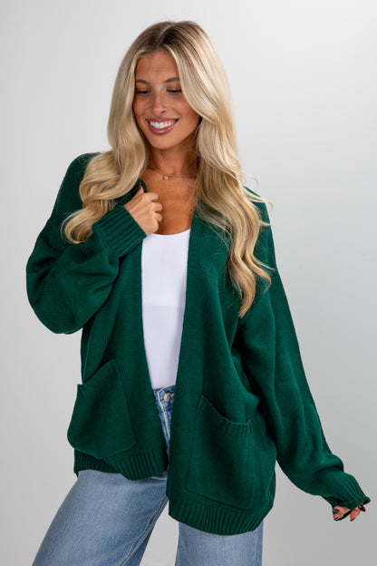 Woman wearing a green cardigan over a white top and blue jeans on a plain background