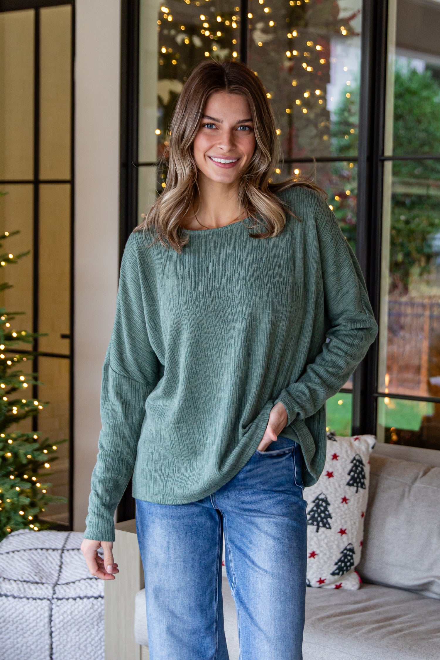 Woman wearing a green sweater and blue jeans standing in a room with festive decorations.