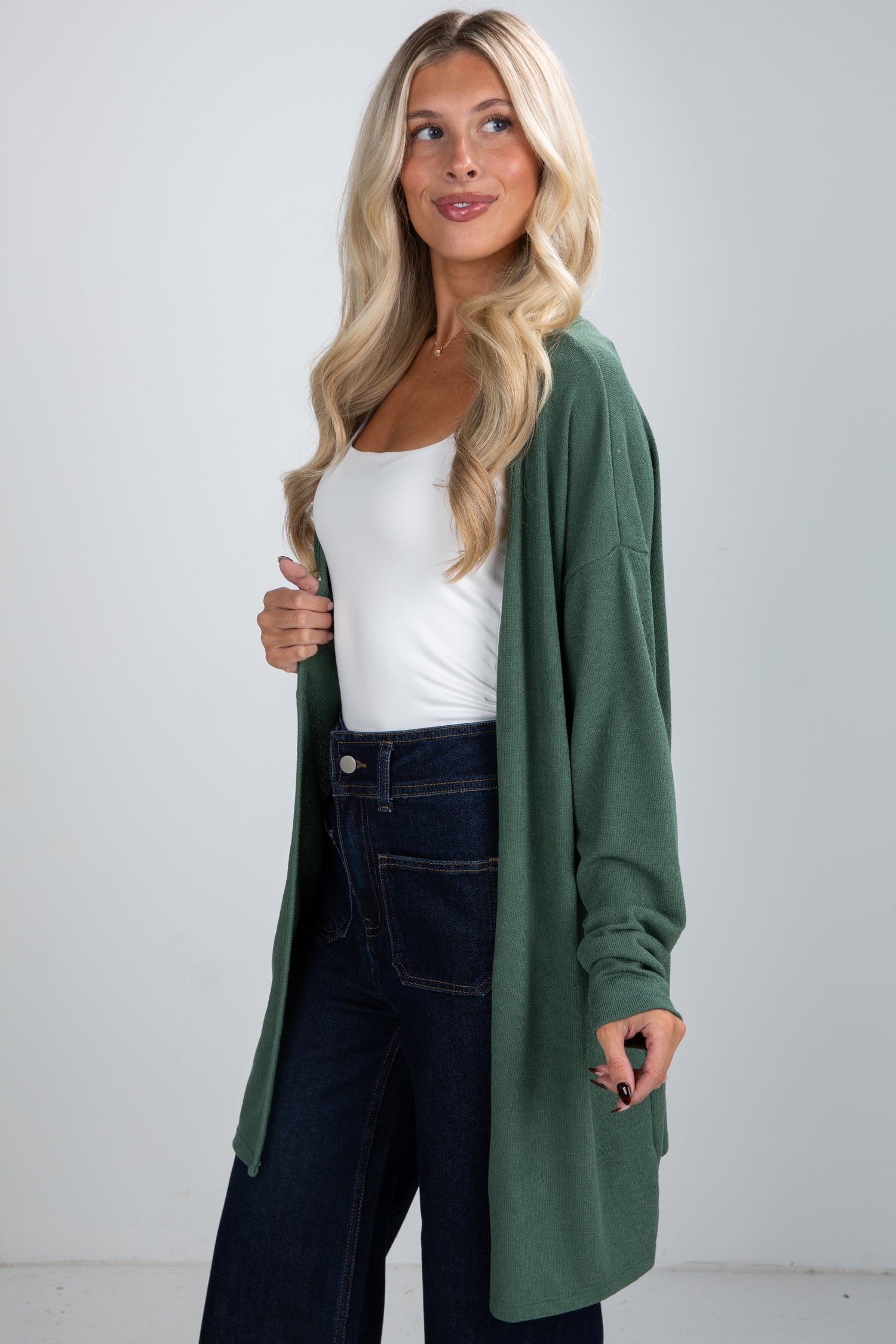 Woman wearing a green cardigan over a white top and dark jeans on a plain background