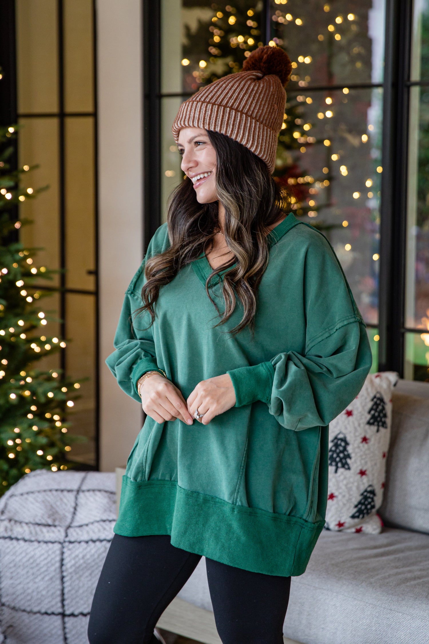 Woman wearing a green sweater and brown beanie in a cozy living room with Christmas decorations.