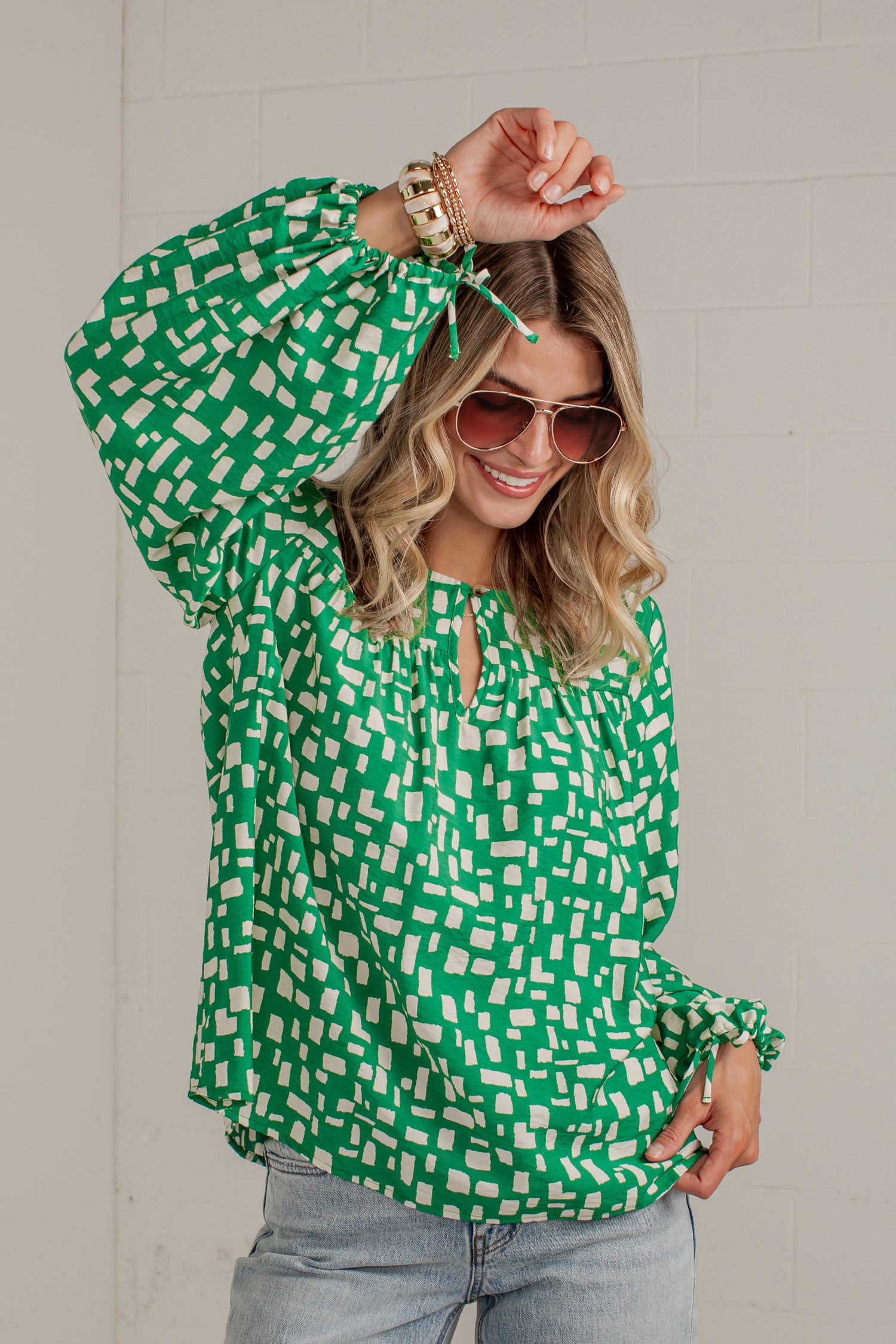 A woman wearing a green and white geometric patterned blouse with long balloon sleeves and a keyhole neckline, paired with light blue jeans and sunglasses, posing against a light gray brick wall.