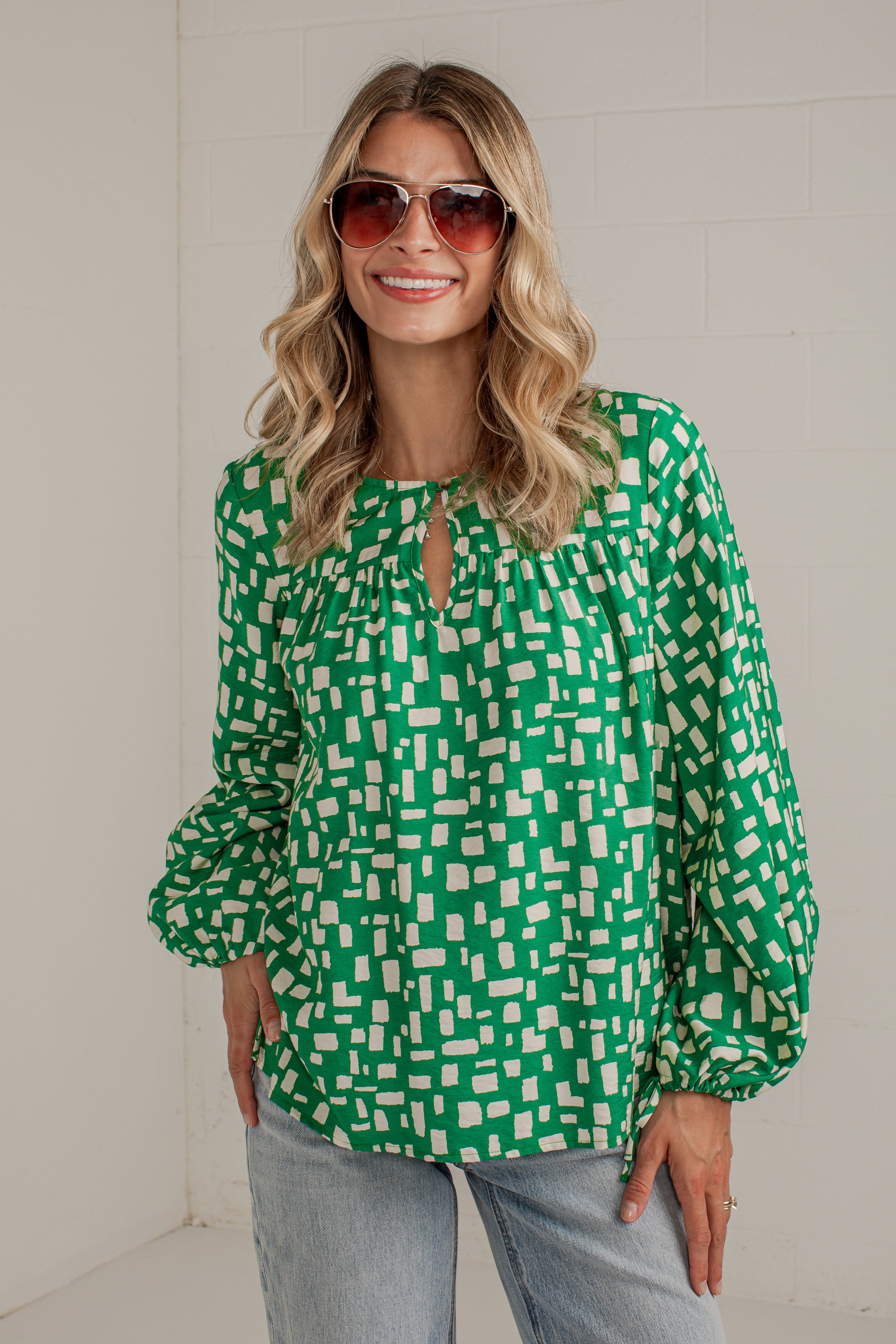 A woman wearing a green and white geometric patterned blouse with long balloon sleeves and a keyhole neckline, paired with light blue jeans and sunglasses, standing against a white brick wall.