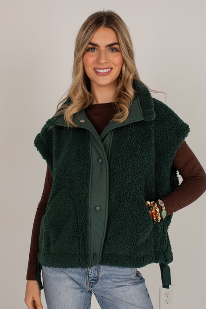 A woman wearing a dark forest green sherpa fleece vest over a brown long-sleeve top and light blue jeans, standing against a plain light gray studio background.