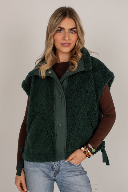 A woman modeling a dark forest green sherpa fleece vest with a button-front closure over a brown long-sleeve top and light-wash blue jeans, set against a plain light gray studio background.