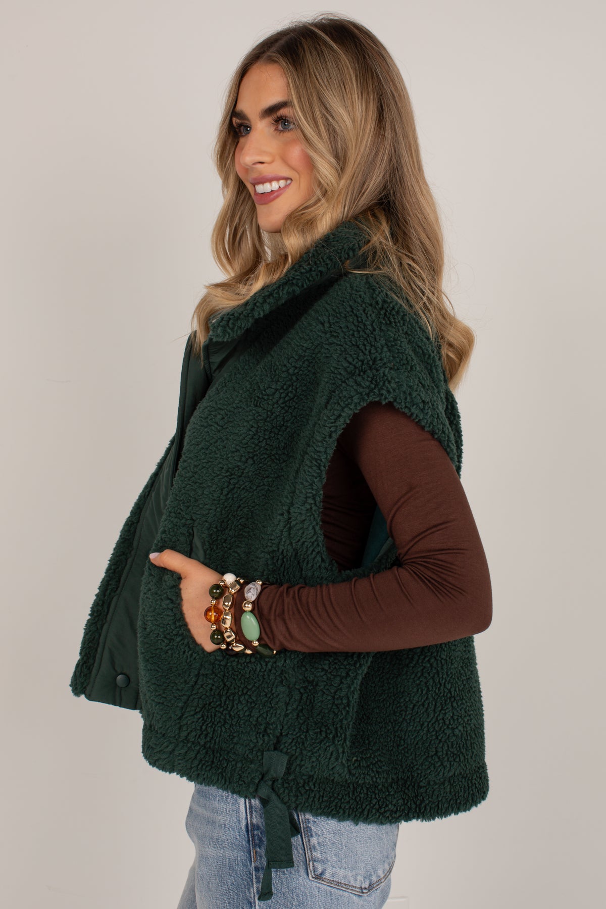 A woman wearing a dark forest green sherpa fleece vest over a brown long-sleeve top and light blue jeans, posing against a neutral light gray background.