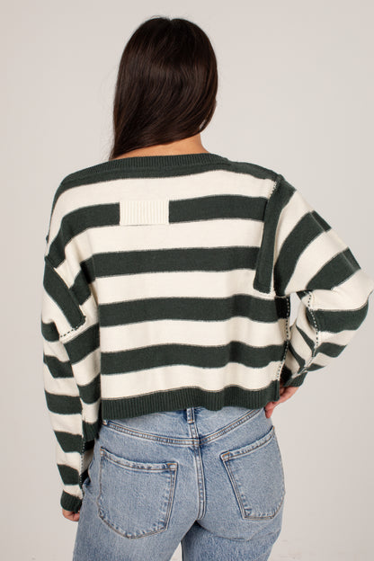 A woman viewed from behind wearing a dark green and off-white striped knit sweater with wide sleeves and light blue jeans, against a plain light gray studio background.