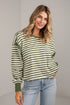 A woman wearing a green and off-white striped long-sleeve top with solid green ribbed cuffs and hem, paired with light-wash blue jeans, standing against a light gray wall.
