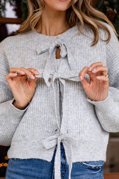 Person wearing a light gray knit cardigan with a tie detail, standing indoors.