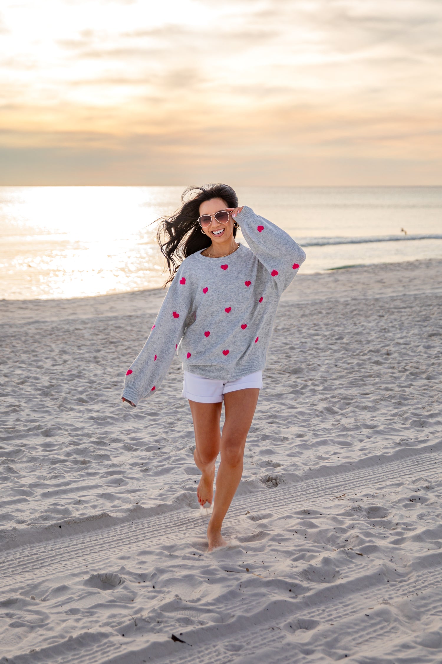 Woman walking on a beach wearing a gray sweater with red heart patterns.