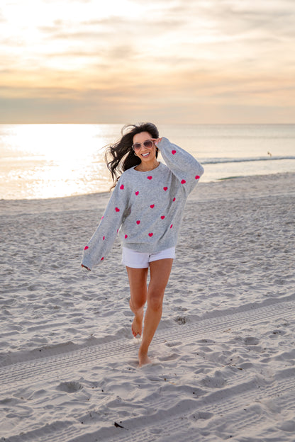 Woman walking on a beach wearing a gray sweater with red heart patterns.