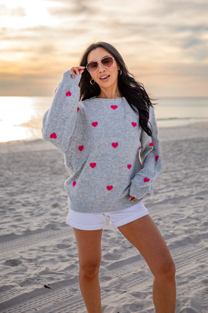 Woman wearing a gray sweater with red heart patterns on a beach at sunset.