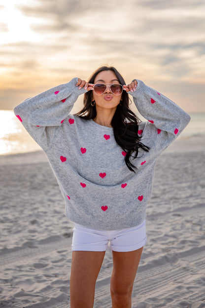 Woman wearing a gray sweater with red heart patterns on a beach at sunset.
