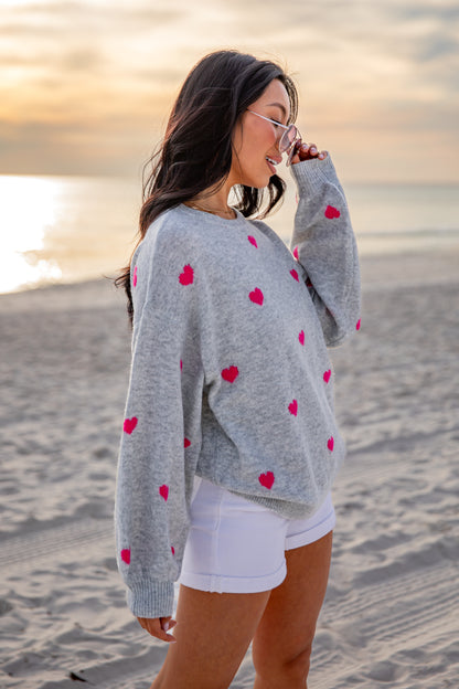 Woman wearing a gray sweater with red heart patterns on a beach at sunset.