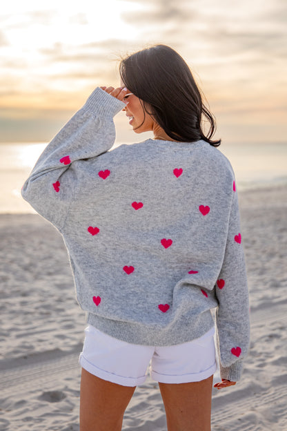 Woman wearing a gray cardigan with red heart patterns on a beach at sunset.