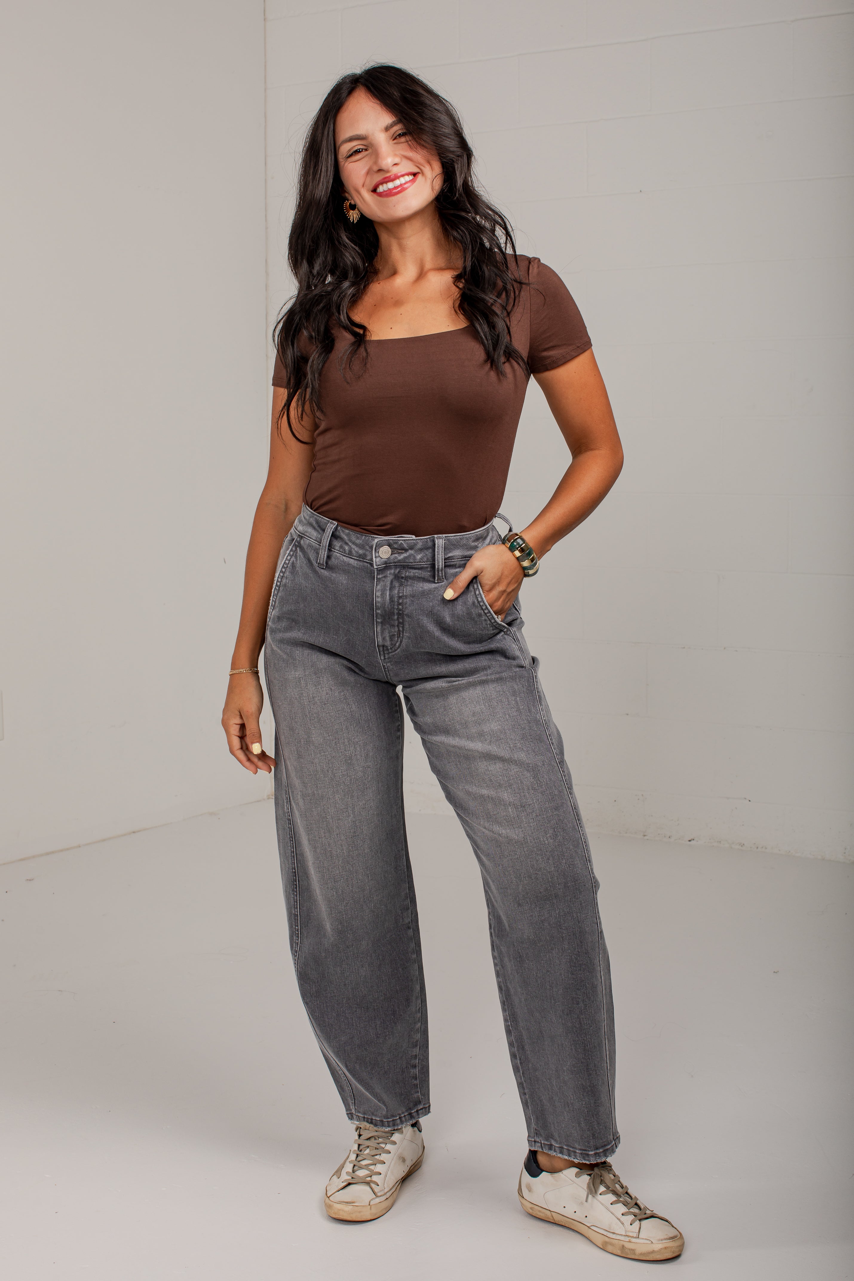 A woman modeling a brown short-sleeve top and gray wide-leg jeans with white sneakers, standing against a plain light gray studio background.