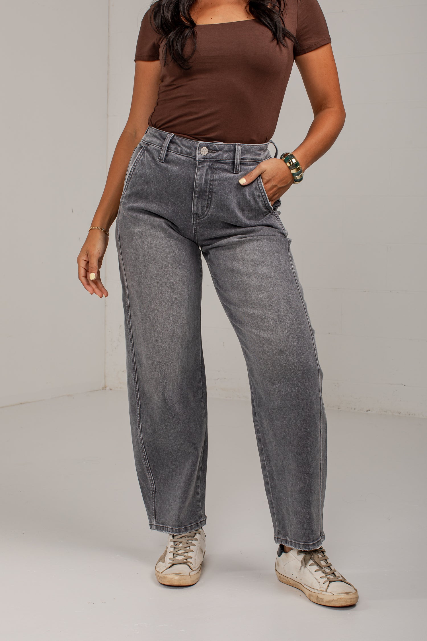 A model wearing a pair of high-waisted, wide-leg gray jeans with a faded wash, styled with a brown scoop-neck t-shirt and beige sneakers against a plain light gray studio background.
