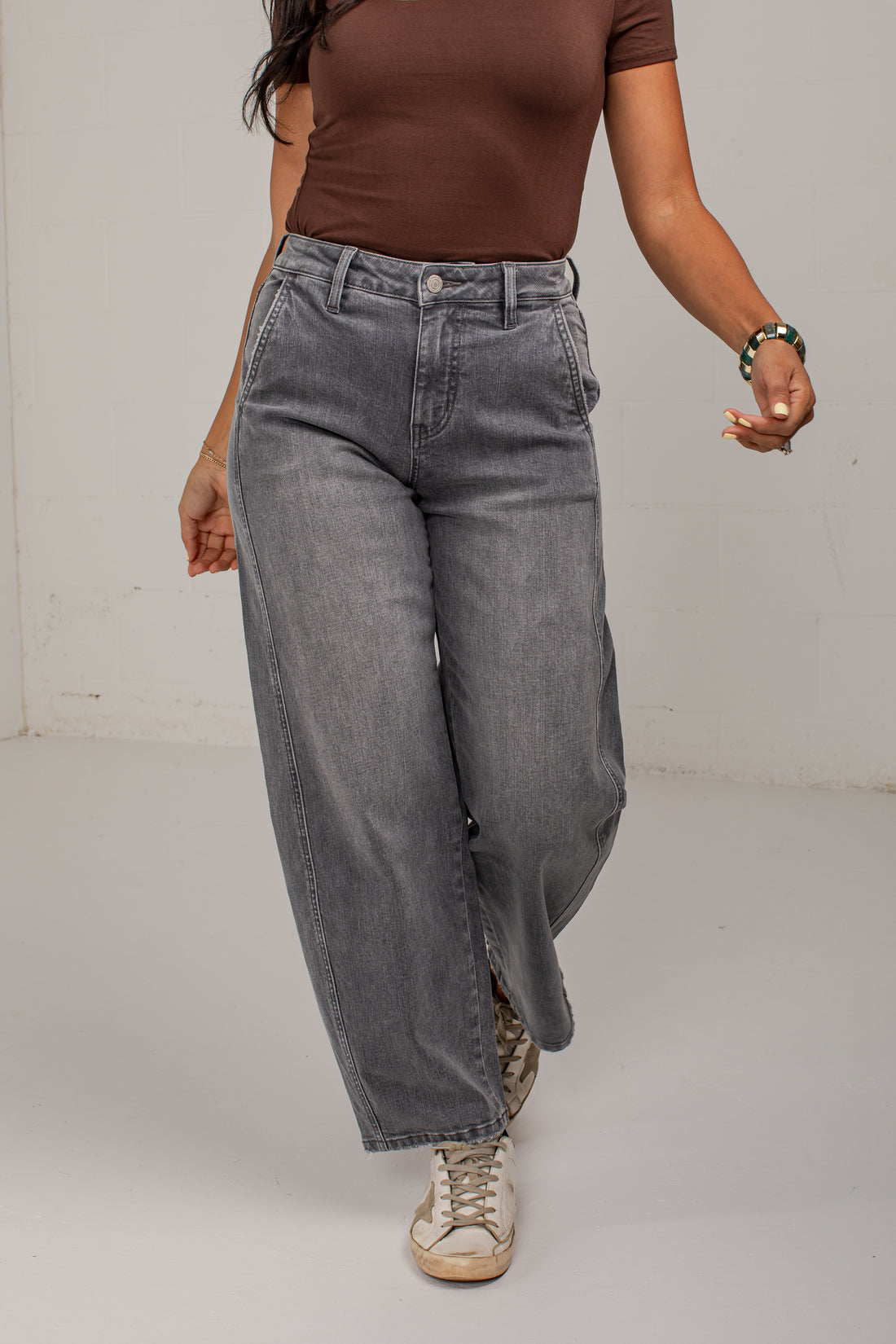 A person wearing wide-leg gray denim jeans with a side slit, paired with a brown short-sleeve top and beige sneakers, shown from the chest down in a studio setting.
