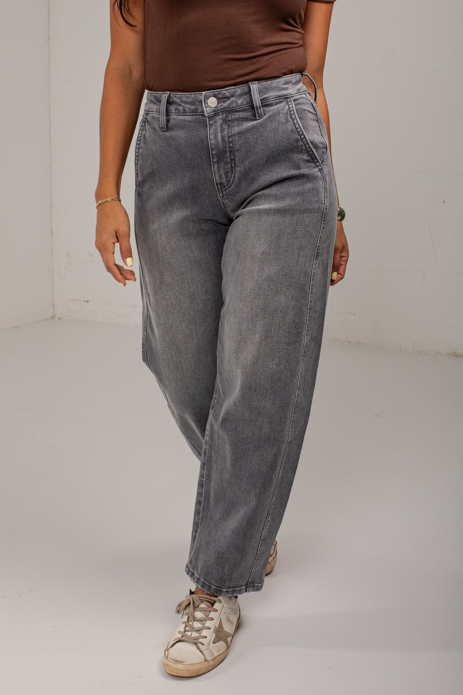 A person wearing loose-fit gray denim jeans with a faded wash, styled with a brown top and beige sneakers, standing in a plain indoor setting.