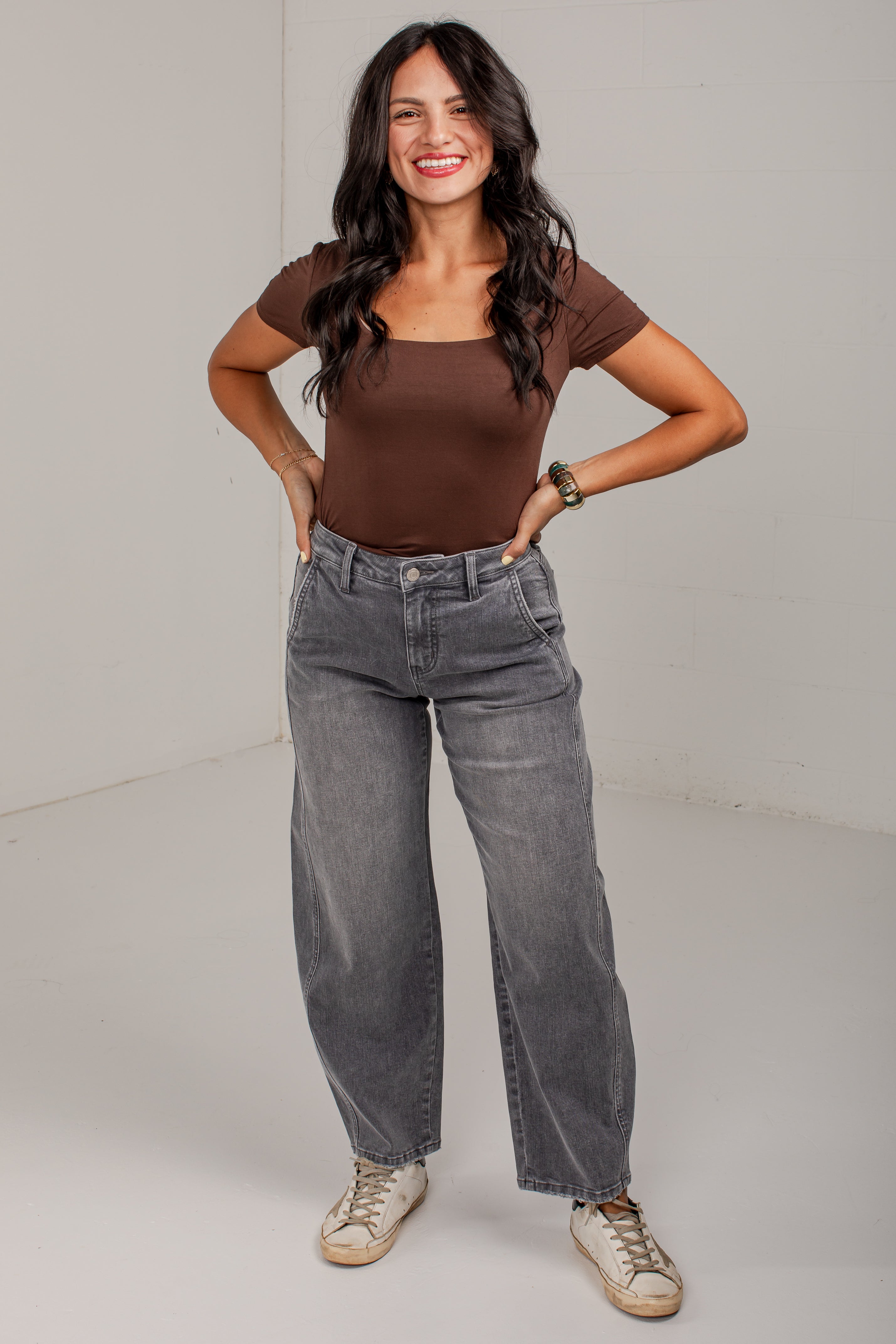 A woman modeling a brown scoop-neck short-sleeve top and gray wide-leg jeans, standing against a plain light gray studio background.