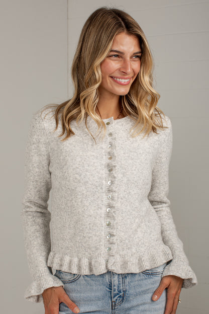 A woman wearing a light heather gray knit cardigan with a button-down front and ruffled hem, paired with blue jeans, against a neutral gray background.