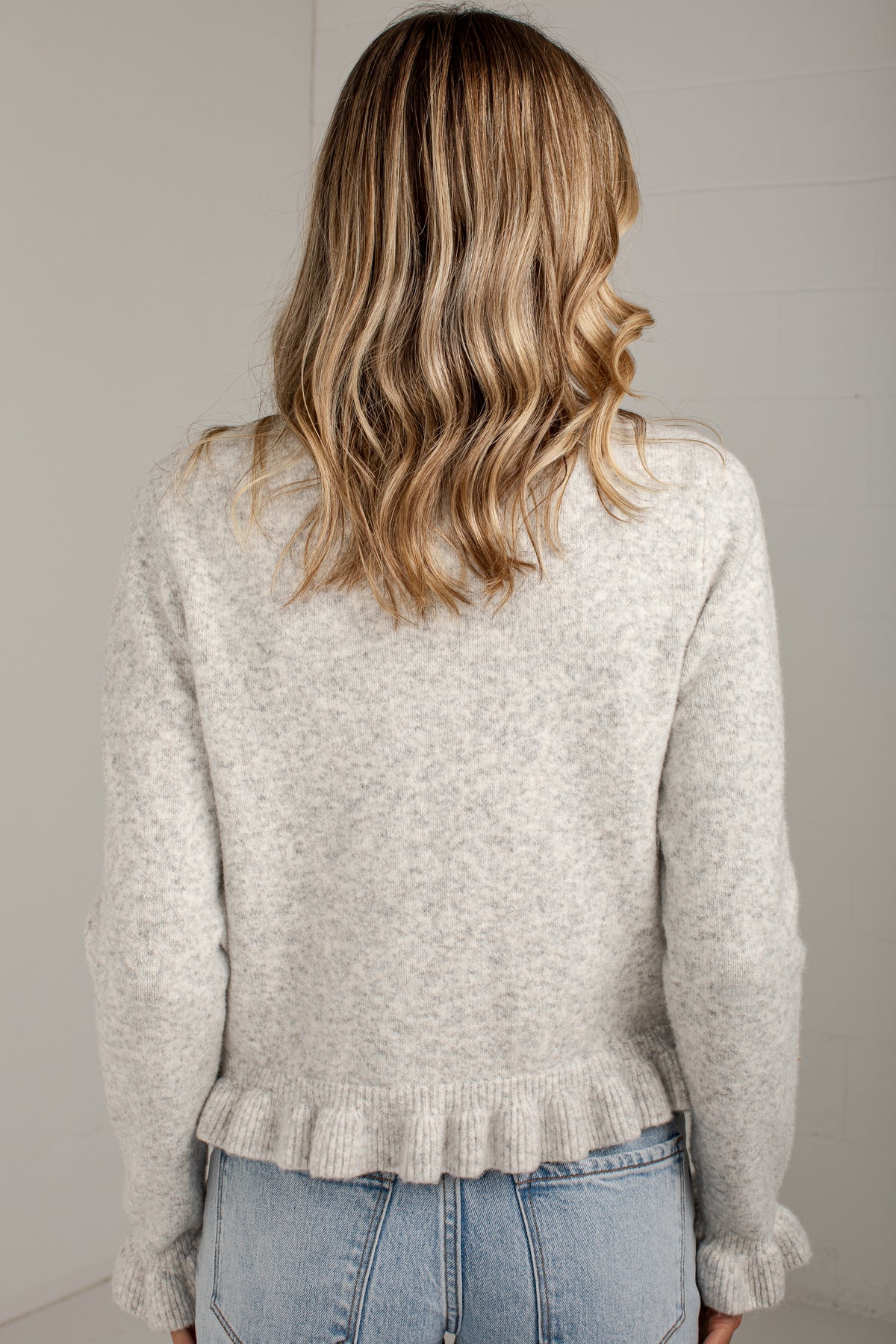 A woman viewed from behind wearing a light heather gray knit sweater with a ruffled hem and light blue jeans, set against a plain light gray wall.