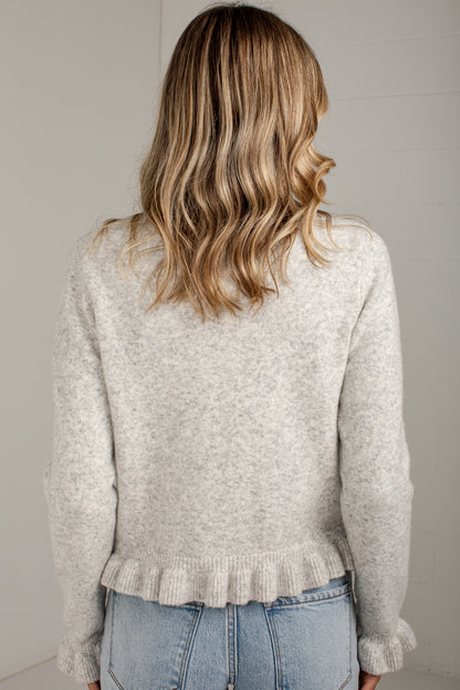 A woman viewed from behind wearing a light heather gray knit sweater with a ruffled hem and light blue jeans, set against a plain light gray wall.