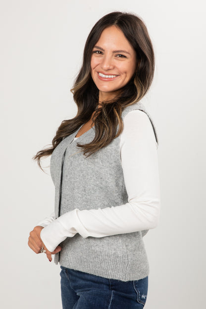 Woman wearing a gray vest over a white long-sleeve shirt on a plain background