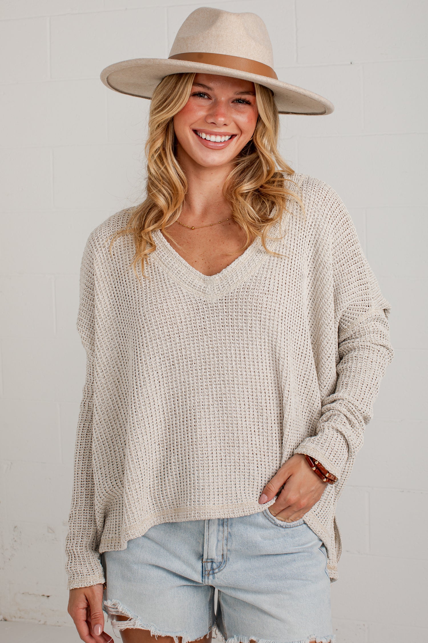 A woman wearing a beige waffle-knit V-neck sweater, a wide-brim beige hat, and light-wash denim shorts, standing against a white brick wall.