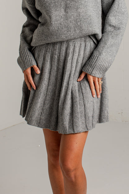 A person wearing a gray ribbed knit sweater and matching pleated mini skirt, standing against a plain light gray wall.