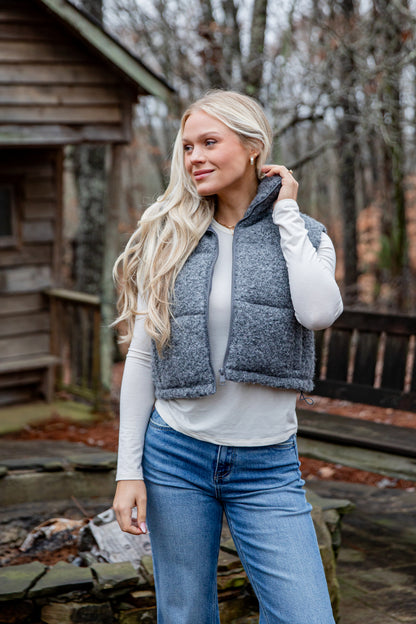 Woman wearing a blue puffer vest and jeans standing outdoors near a wooden cabin.