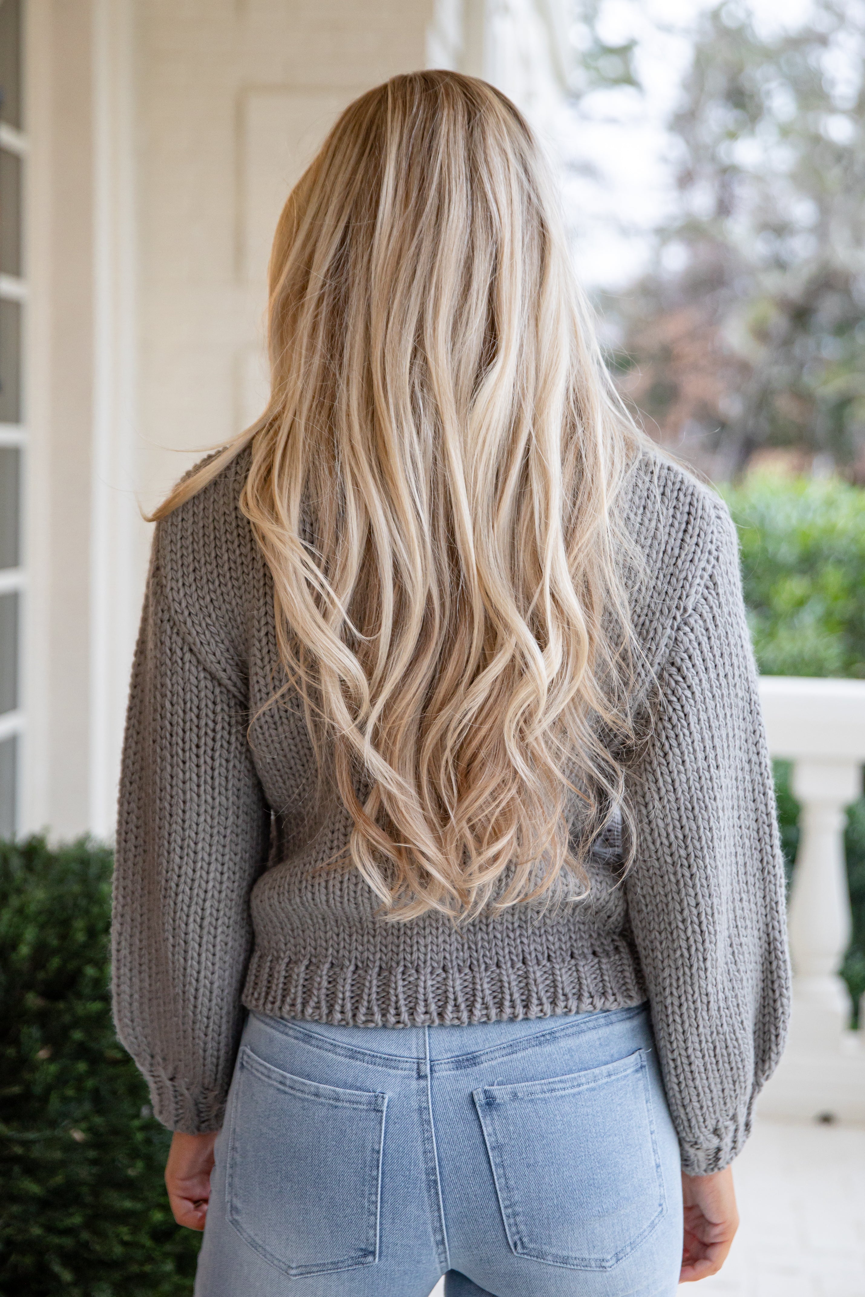 Person with long blonde hair wearing a gray sweater and light blue jeans outdoors.