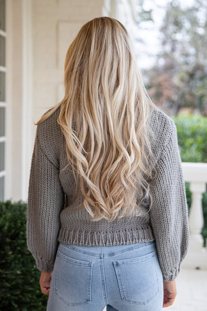 Person with long blonde hair wearing a gray sweater and light blue jeans outdoors.
