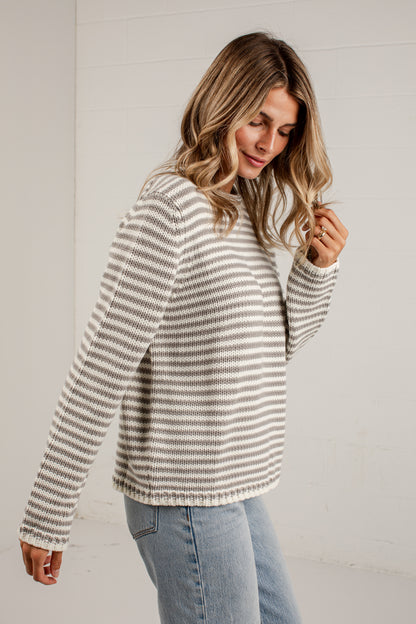 A woman wearing a beige and white horizontally striped knit sweater and light blue jeans, standing against a white paneled wall.