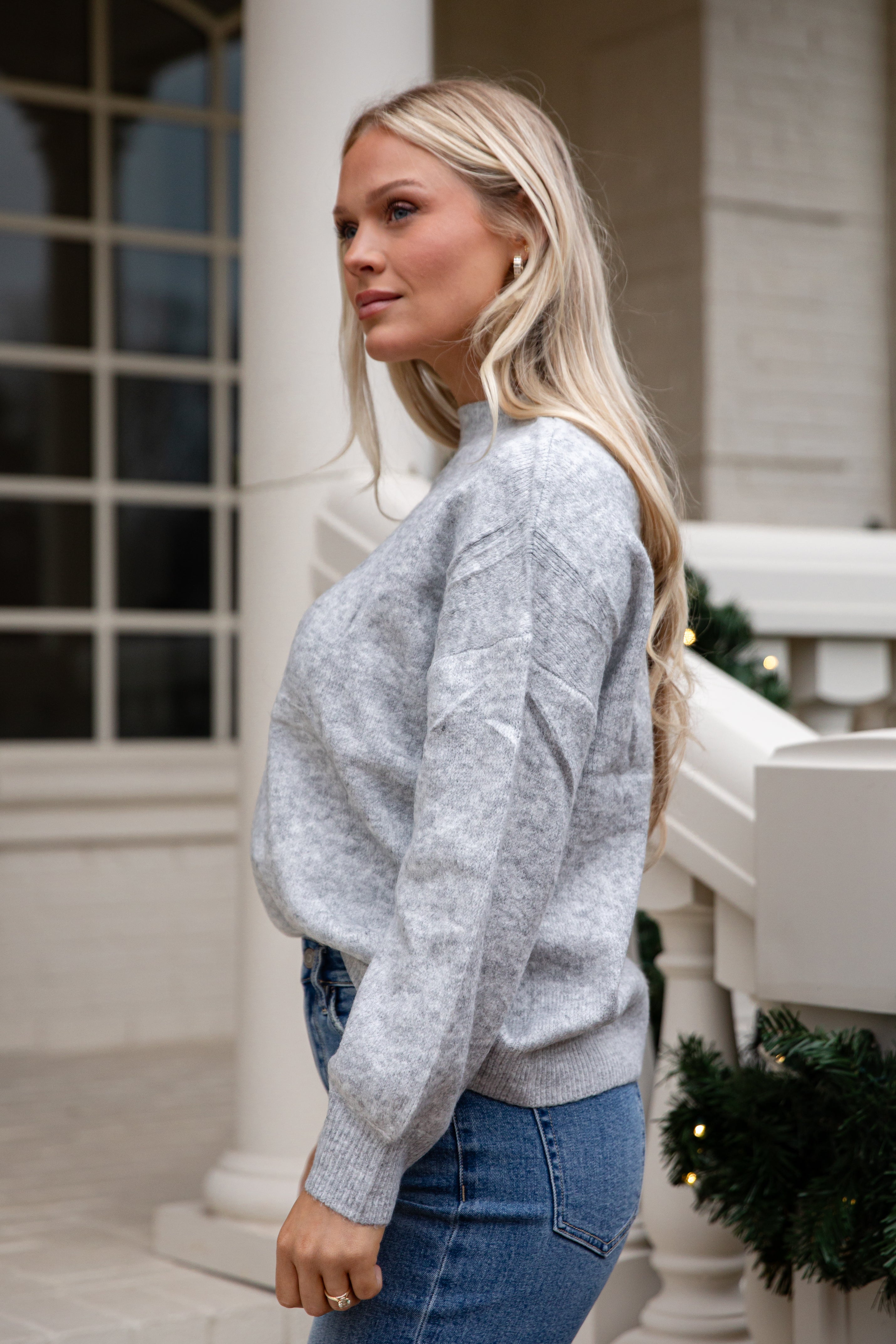 Woman wearing a gray sweater and blue jeans standing on a porch.