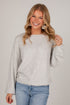 A smiling blonde woman wearing a light heather gray long-sleeve knit sweater and blue jeans, standing against a plain light gray studio background.