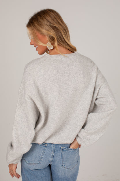 A woman shown from the back wearing a light heather gray knit sweater and blue jeans against a plain light gray studio background.